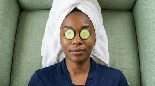 Mulher negra reclinada em um sofá verde, com toalha branca enrolada no cabelo e fatias de pepino sobre os olhos, em momento de relaxamento e autocuidado.