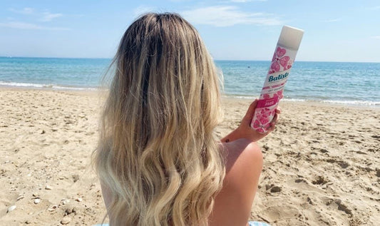 Mulher loira de costas na praia segurando um shampoo a seco batiste